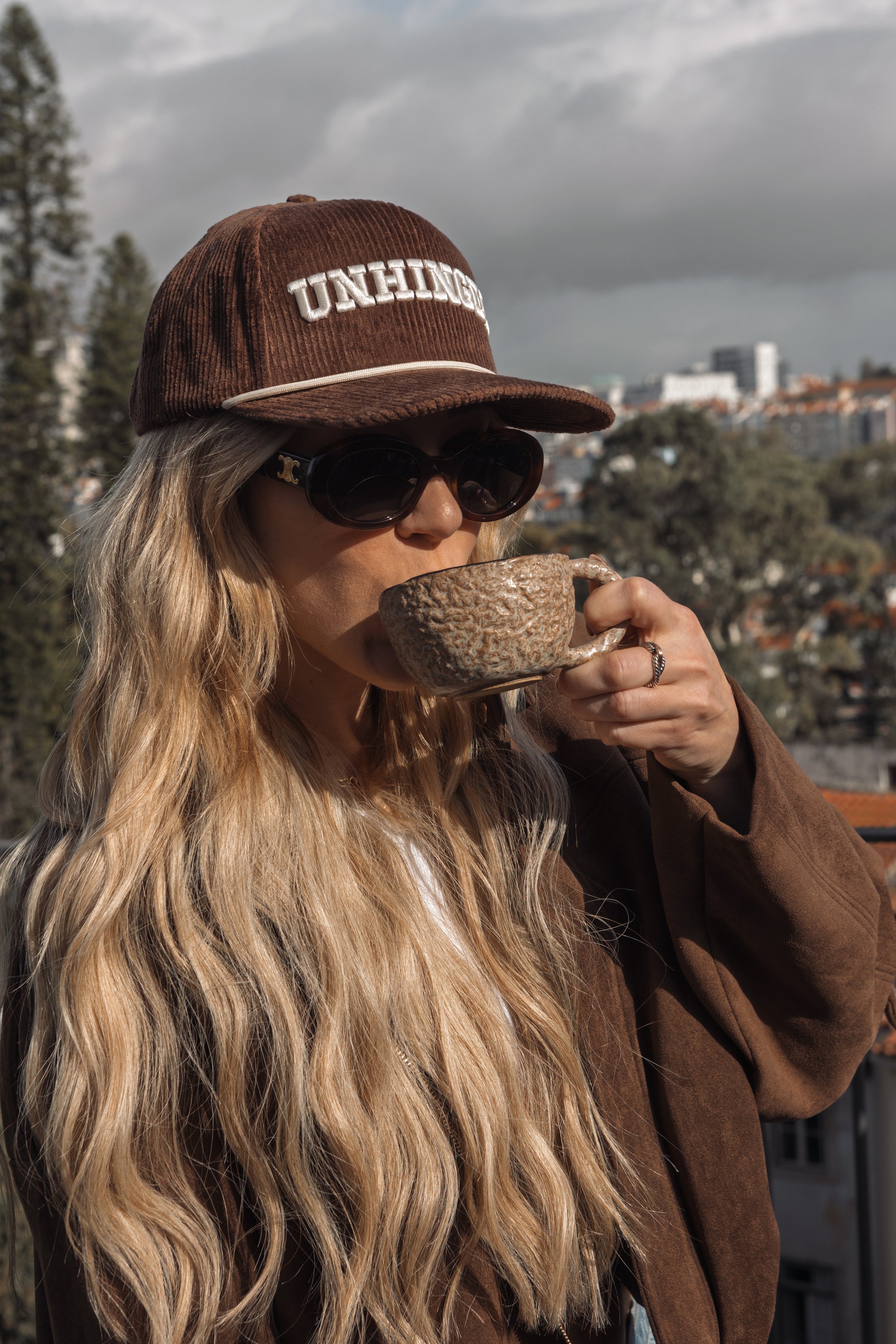 Person wearing a brown 'UNHOKED' cap and sunglasses, drinking from a textured mug with a cityscape in the background.