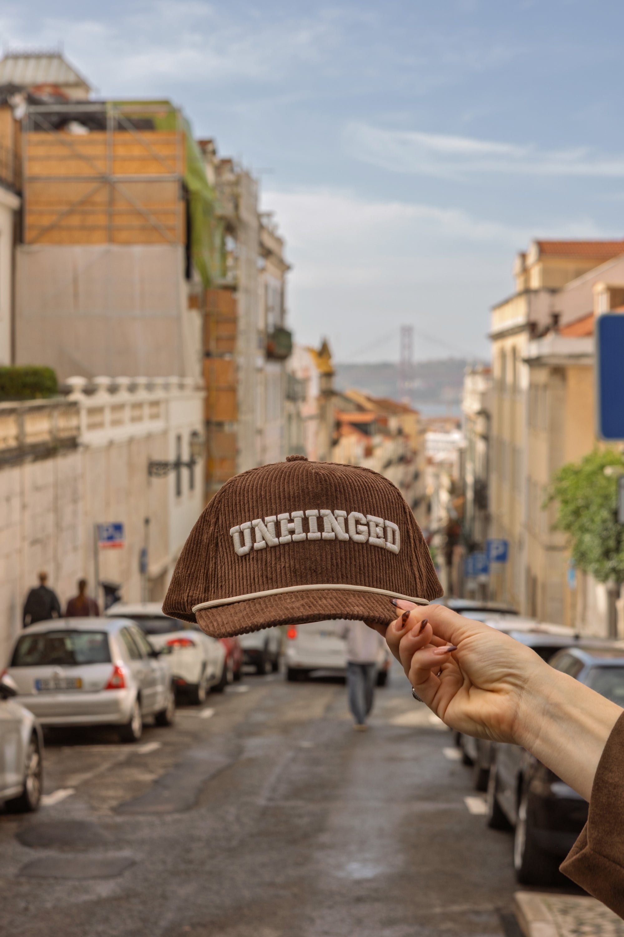 Brown cap with 'UNHINGED' text held up in front of a city street.