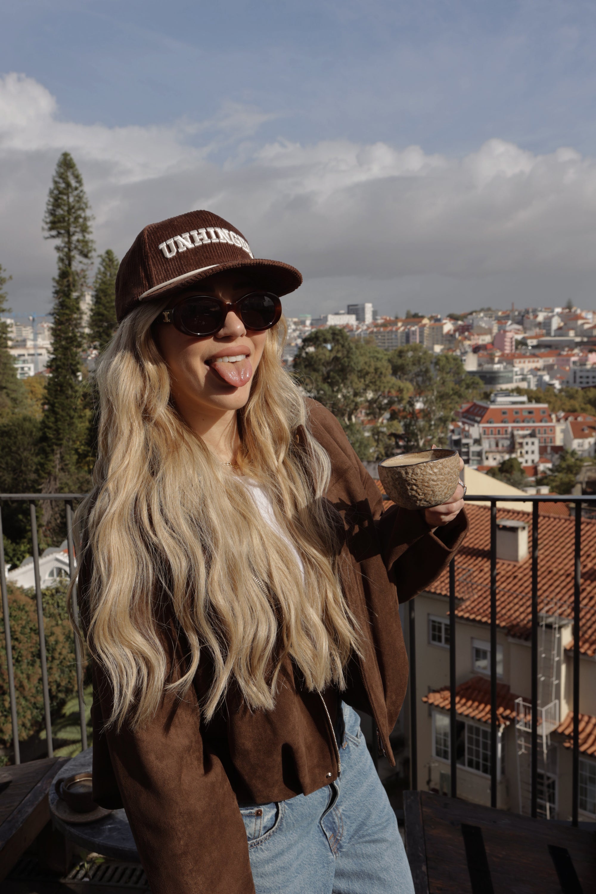 Person with long blonde hair wearing a brown hat and coat, standing outdoors with a cityscape in the background.